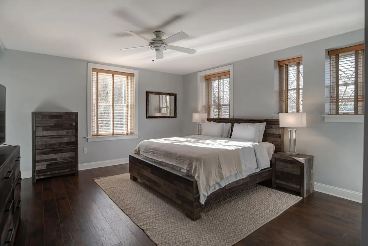A tranquil bedroom featuring a plush king bed with crisp linens, flanked by matching nightstands, a large mirror reflecting soft natural light, and expansive windows bringing in the morning sun.