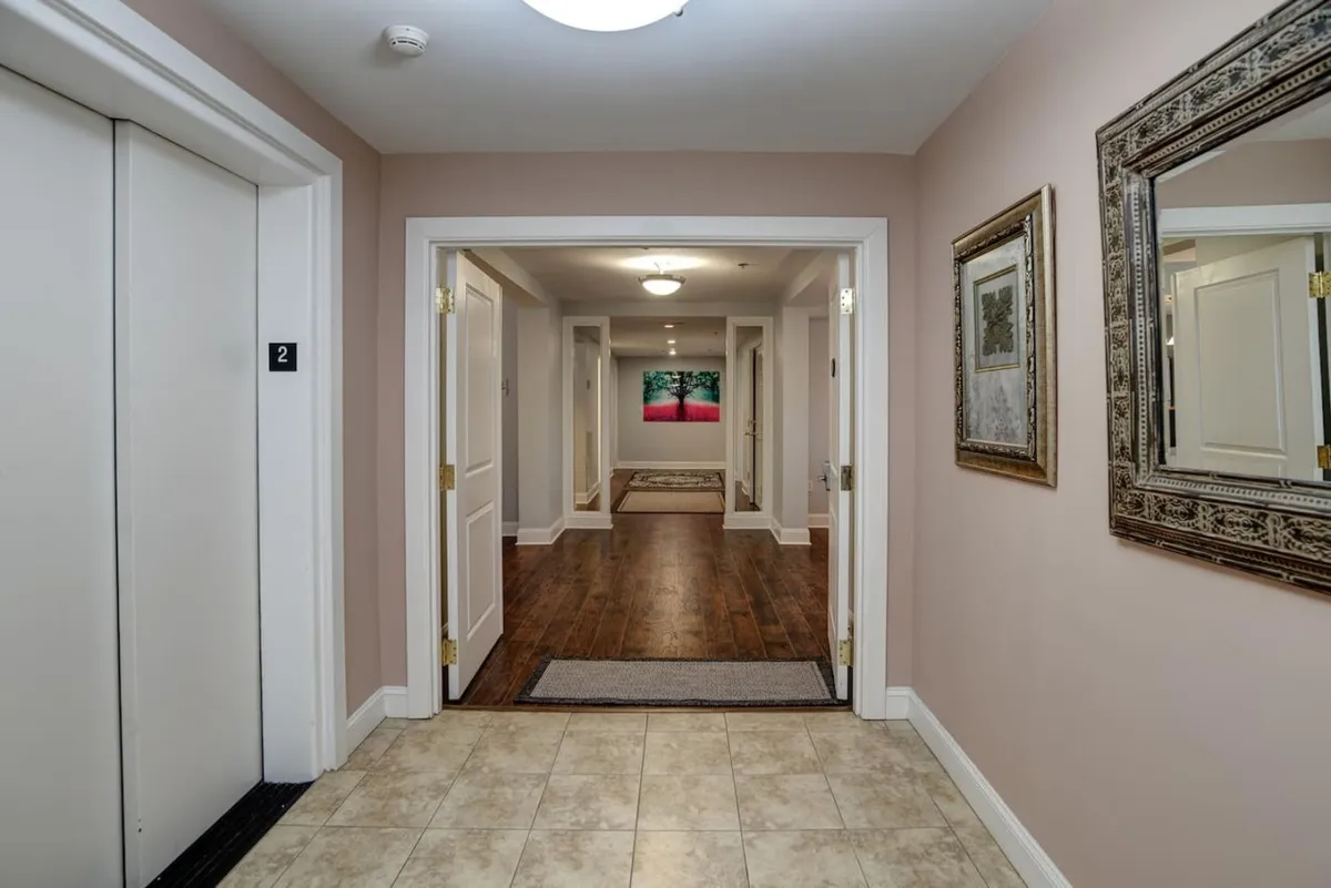A refined hallway scene with elevator doors, art displayed on the wall, a decorative mirror, and a polished wooden floor. The setting is inviting, suggesting easy movement and a welcoming first impression.