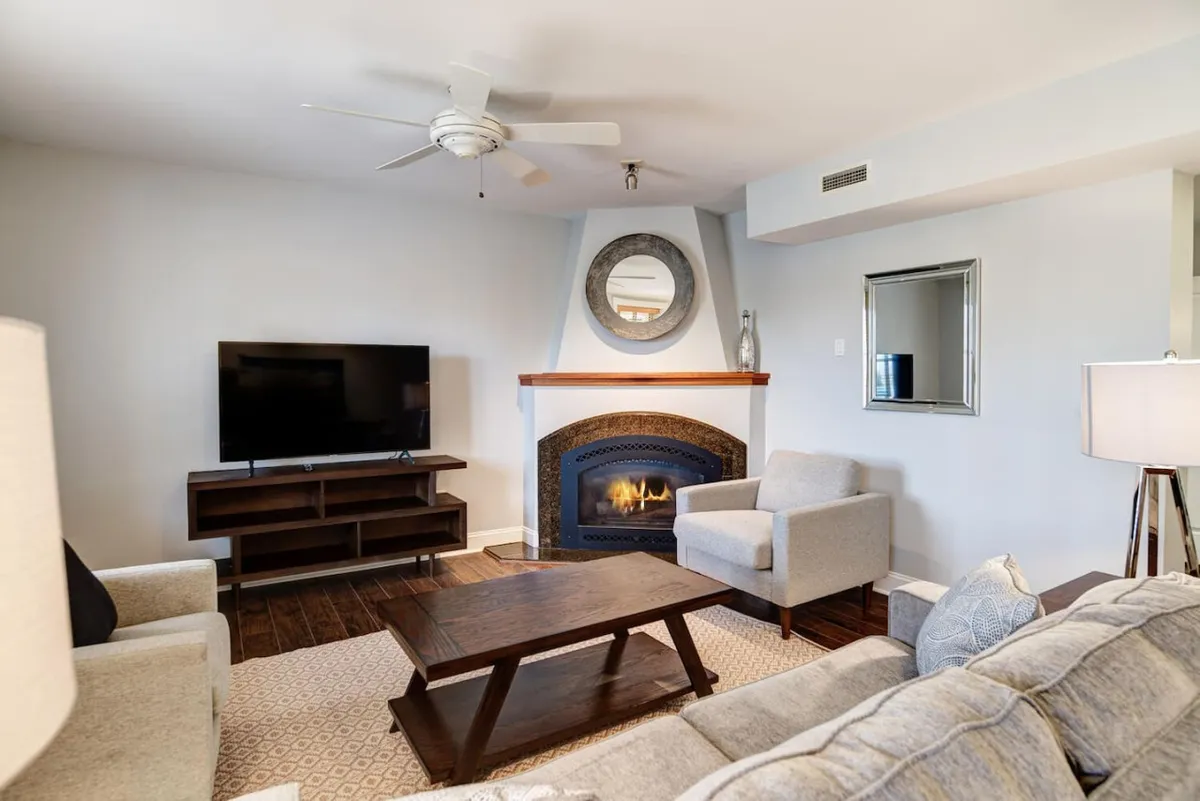 A cozy living room featuring a plush sofa, a coffee table, a modern fireplace, and a TV. Gentle light streams in, creating a warm and inviting atmosphere perfect for relaxing or socializing with friends and family.