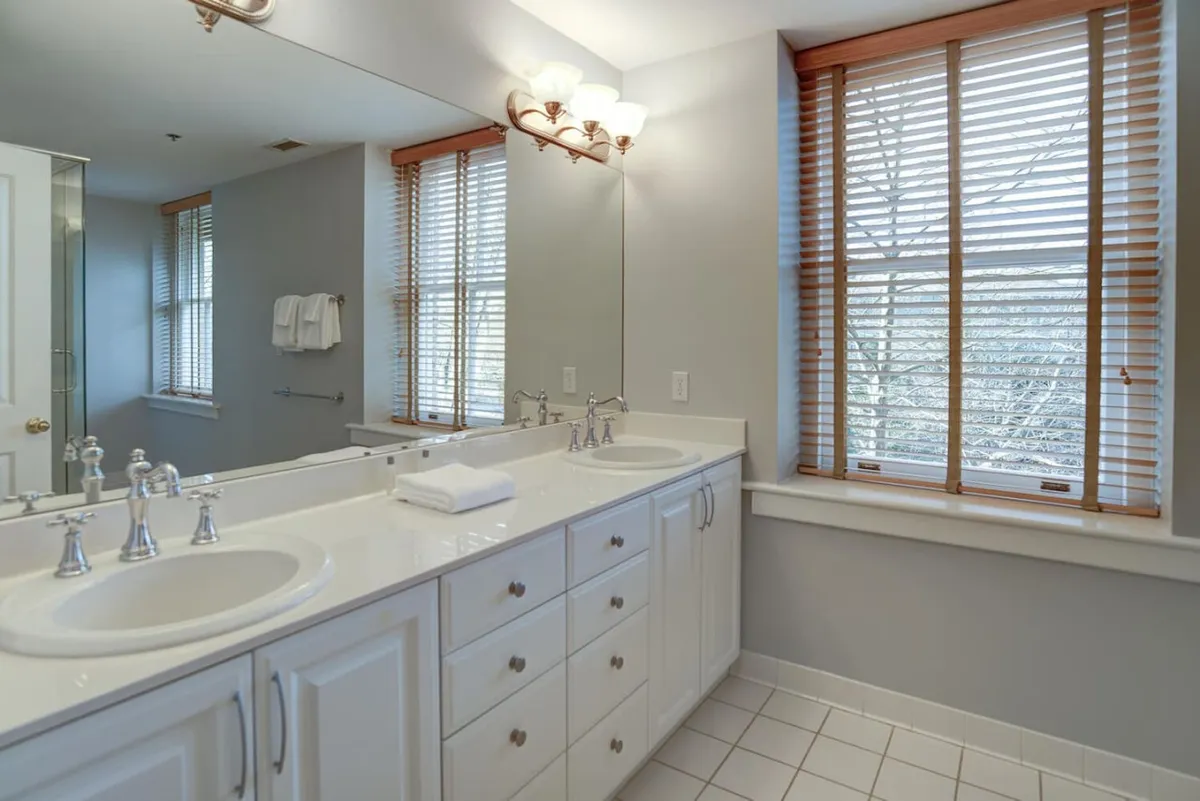 A bright bathroom with double sinks, a large mirror, and natural light pouring in from windows adorned with wooden blinds—offering a tranquil space for morning routines and moments of reflection.