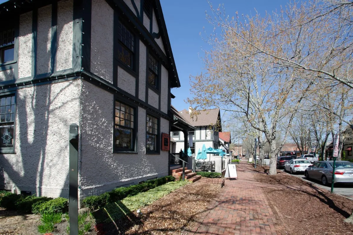 This exterior shot showcases the building's distinctive architecture, with a welcoming sidewalk leading up to the entrance and leafy trees lining the street. The visible outdoor cafes and nearby shops hint at the lively, accessible neighborhood atmosphere.
