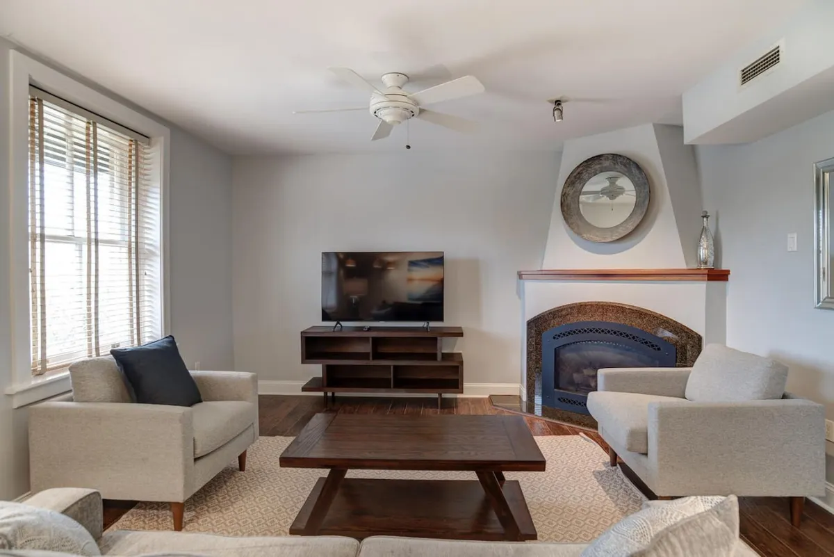 This image showcases a spacious living room featuring a plush sofa, coffee table, television, and a cozy fireplace. Ample natural light pours in through large windows, illuminating the hardwood floors and highlighting tasteful, historic-inspired décor. The inviting space hints at both relaxation and social gatherings.