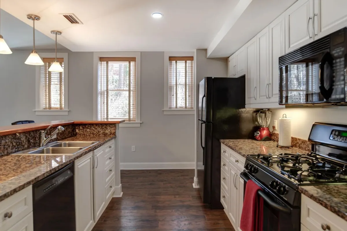 This image captures a renovated kitchen with gleaming granite countertops, modern stainless steel appliances, warm wooden cabinets, and sunlight streaming through ample windows. The open layout invites conversation and social gatherings, while the countertops and appliances highlight the kitchen's functionality and style.