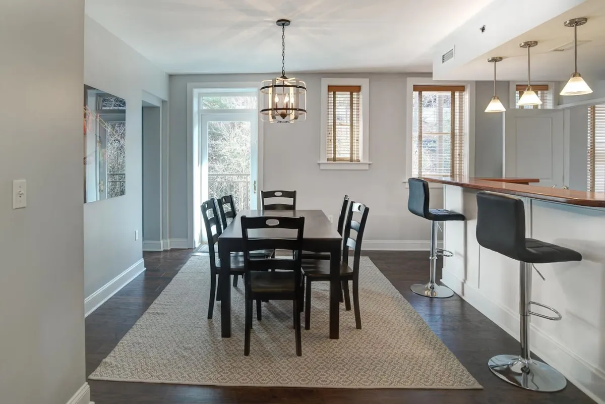 A bright dining area with a substantial dining table, stylish stools at a kitchen bar, a striking chandelier, and large windows that bathe the space in sunlight. The hardwood floors and elegant touches evoke the condo's historic character while supporting group gatherings and family dinners.